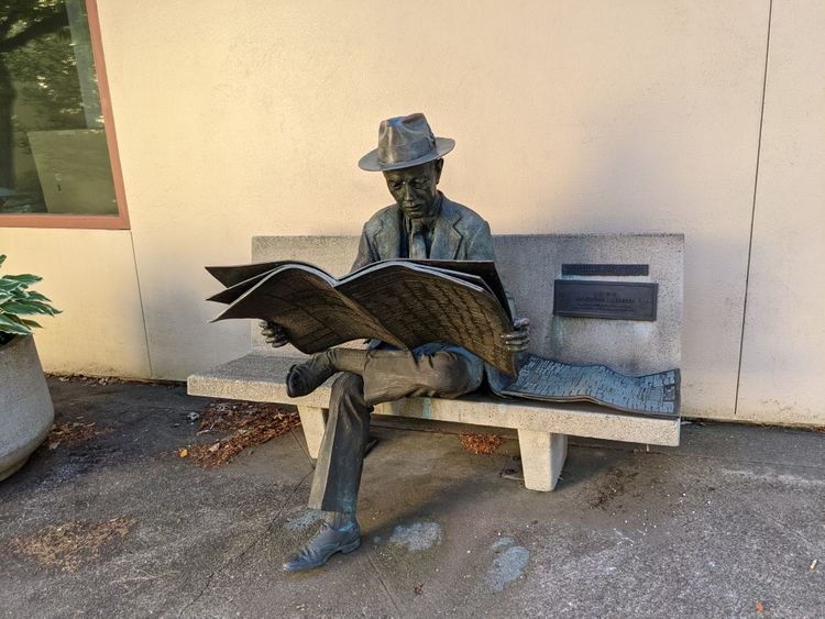 A weathered statue of a man in a suit and h sitting on a cement bench reading a newspaper